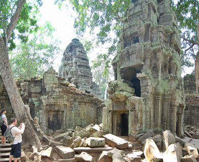 Ancient stone temple towers covered in green moss with large trees growing through the ruins, scattered stone blocks on the ground, and two tourists photographing on the left side