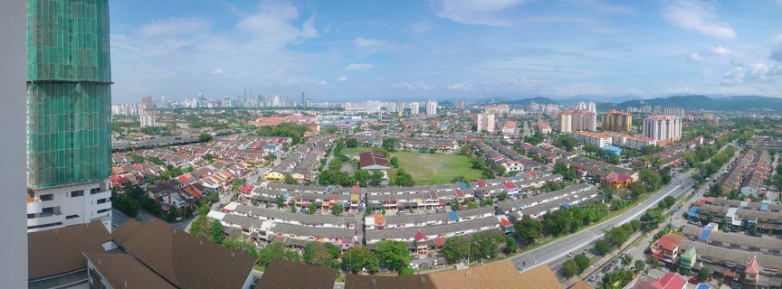 Aerial view of Singapore showing residential housing estates with varied roof colors, a building under construction with green safety netting, a green sports field, major roadway, and distant city skyscrapers under a blue sky with scattered clouds