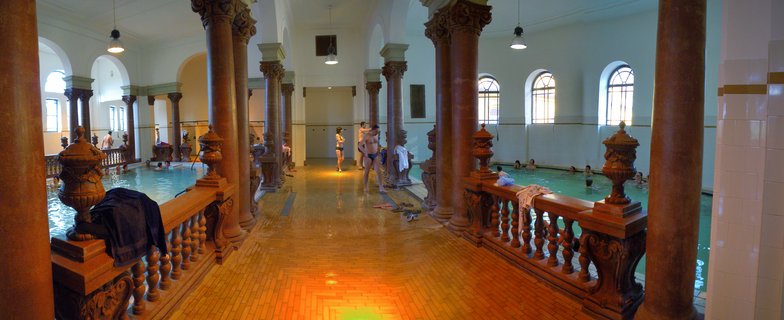 Interior of ornate thermal bath with red marble columns, two swimming pools with people bathing, arched windows, and tiled floors in Budapest