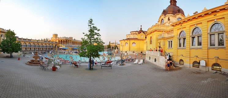 Wide view of Széchenyi Thermal Bath in Budapest showing yellow Neo-Baroque buildings, outdoor thermal pools with swimmers, courtyard with fountains and benches, and visitors enjoying the spa facilities during golden hour