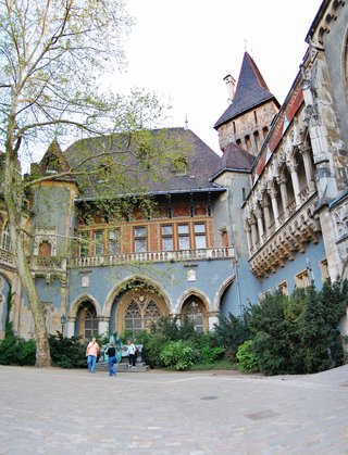 Historic castle courtyard in Budapest featuring Gothic Revival architecture with arched windows, ornate stone balconies, red tile roofs, and visitors walking on cobblestone pavement beneath spring trees