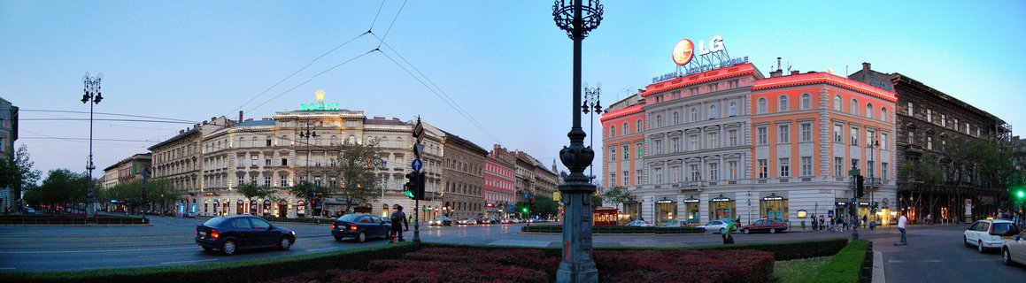 Wide street view of Ferenciek tere square in Budapest at dusk showing illuminated historic buildings, ornate street lamps, cars on the road, pedestrians, and commercial signage against a blue twilight sky