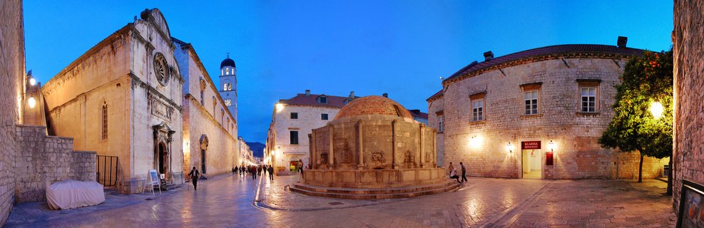Historic stone fountain illuminated at twilight in Dubrovnik's Old Town square with Franciscan Monastery and bell tower, people walking on cobblestone plaza