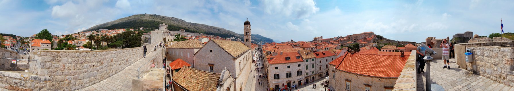 Panoramic photograph showing Dubrovnik's historic Old Town with orange-red tiled roofs, stone buildings, a bell tower, and tourists walking on ancient city walls with a green hillside in the background under a partly cloudy blue sky