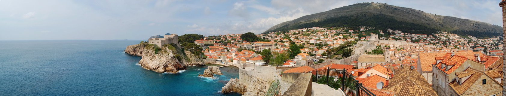 Aerial view of Dubrovnik's historic Old Town showing red terracotta rooftops, stone city walls, Fort Lovrijenac on rocky coastline, turquoise Adriatic Sea, and forested Mount Srđ in the background under a partly cloudy sky