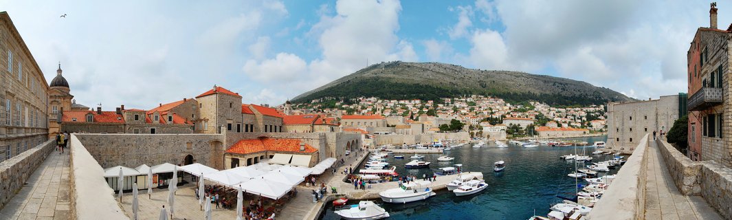 Panoramic view of Dubrovnik harbor with white boats in blue water, surrounded by historic stone buildings with orange-red roofs, city walls in foreground, mountain in background under partly cloudy sky