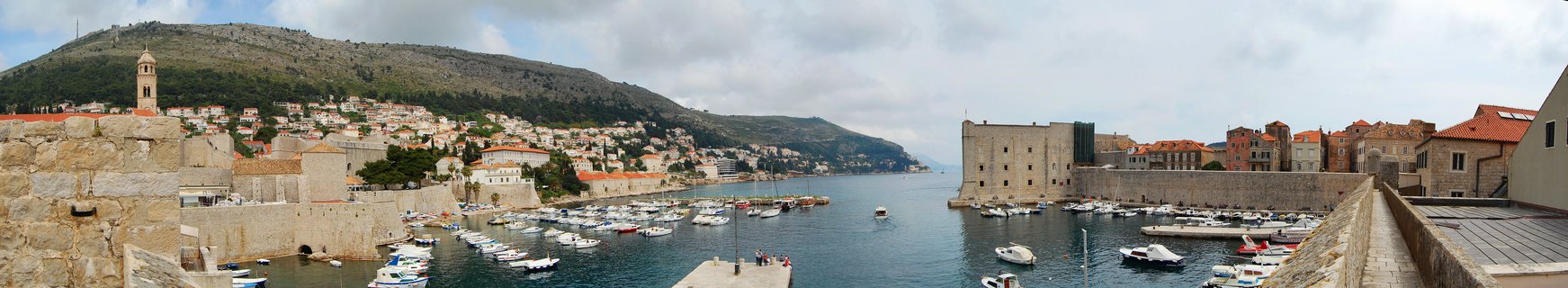 Panoramic view of Dubrovnik's Old Harbor showing medieval stone city walls, fortifications, boats in the water, red-roofed buildings on hillside, and Mount Srđ in the background