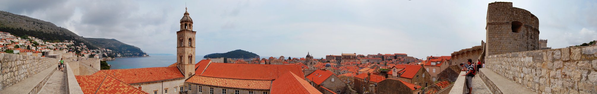Panoramic photograph showing Dubrovnik's Old Town from the city walls with red terracotta rooftops, a prominent bell tower, stone fortifications, and the Adriatic Sea visible in the distance under an overcast sky.