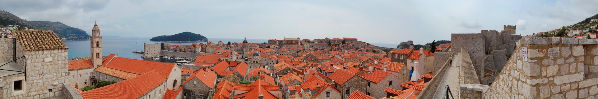 Panoramic photograph showing Dubrovnik's historic Old Town with orange-red tiled rooftops, stone buildings, a bell tower, medieval fortress walls, and the Adriatic Sea with an island in the background under cloudy skies