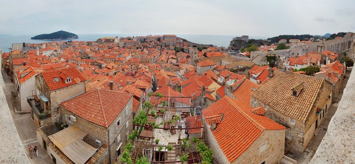 Panoramic aerial view of Dubrovnik Old Town showing dense cluster of orange terracotta rooftops on stone buildings, with Adriatic Sea and green island in background under cloudy sky
