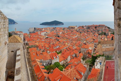 Aerial view of Dubrovnik Old Town showing dense orange terracotta rooftops, stone buildings, church towers, with blue Adriatic Sea and green Lokrum Island in background