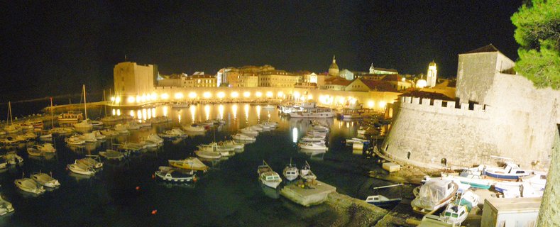 Night photograph of Dubrovnik's Old Harbor showing illuminated medieval stone walls, numerous moored boats in dark water, and historic buildings with a cathedral dome visible in the background