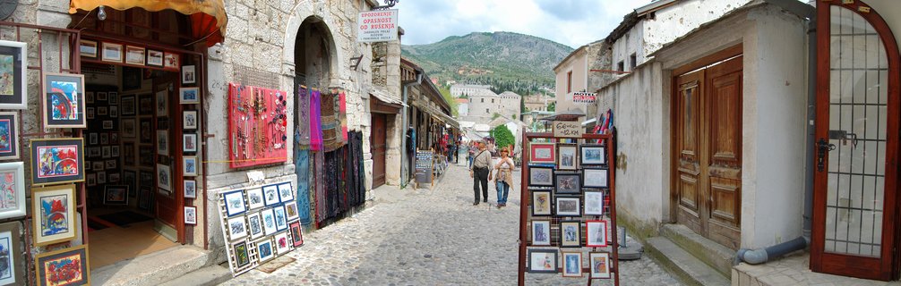 Wide cobblestone street in Mostar Old Town with stone buildings, artisan shops displaying colorful textiles and framed paintings, tourists walking, hills with buildings in background