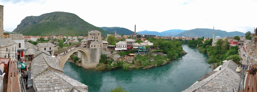 Panoramic view of Stari Most (Old Bridge), a 16th-century Ottoman stone arch bridge spanning the turquoise Neretva River in Mostar, Bosnia and Herzegovina. Historic stone buildings with red terracotta roofs line the riverbank, multiple mosque minarets rise above the Old Town, and forested Velež mountain forms the backdrop under an overcast sky. Tourists walk across the bridge.
