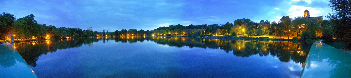 Panoramic view of a lake at twilight with illuminated trees along the far shore, their lights reflecting in the calm blue water, and a church tower visible on the right side