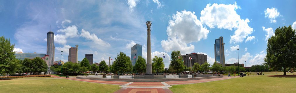Panoramic view of Centennial Olympic Park in Atlanta showing a tall stone column fountain, green lawns, trees, and downtown skyscrapers under a blue sky with white clouds