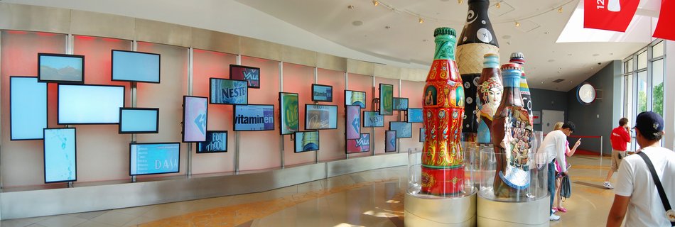 Museum interior with large decorative Coca-Cola bottles painted with cultural designs, visitors walking past a wall of television screens displaying brand content