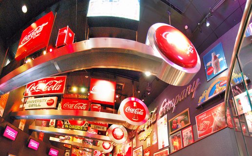 Museum display featuring multiple red Coca-Cola signs, vintage advertising memorabilia, and illuminated brand logos arranged on curved metallic structures with framed historical posters on walls