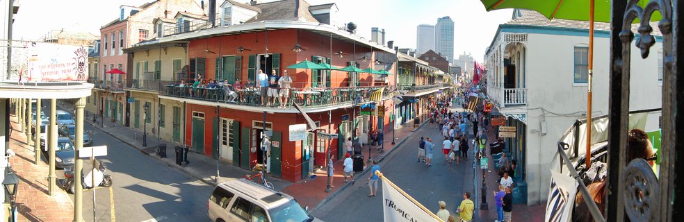 Elevated view of a busy French Quarter street in New Orleans showing historic buildings with balconies, pedestrians walking below, and outdoor dining areas with green umbrellas