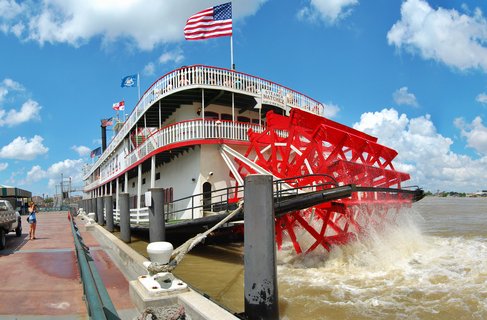 A white multi-deck paddlewheel steamboat with a large red paddlewheel churning brown river water, docked at a concrete pier under blue sky with American flag flying