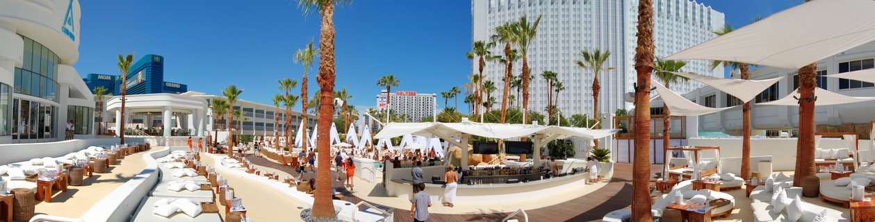 Panoramic view of a luxury resort pool deck with white cabanas, palm trees, shade sails, and hotel buildings under a clear blue sky in Las Vegas