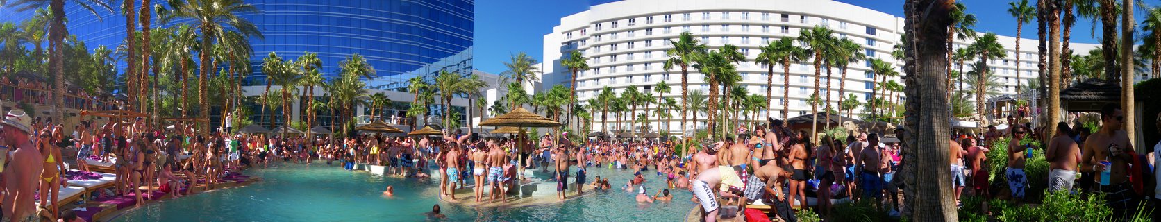 Large crowd of people at a Las Vegas resort swimming pool with hotel buildings and palm trees under blue sky