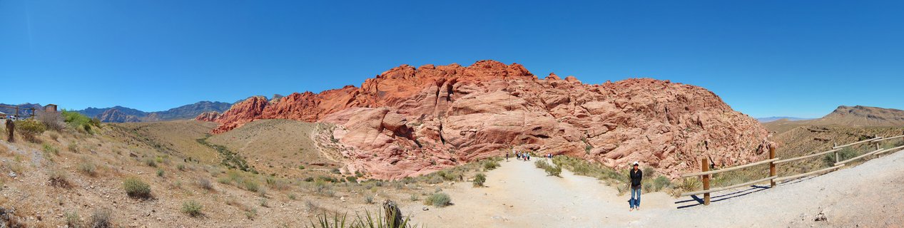 Wide desert landscape showing red sandstone rock formations under clear blue sky with hiking trail, wooden fence, and small figures of people walking toward the rocks