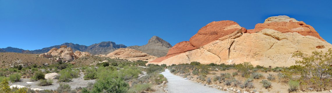 Desert landscape showing red and white sandstone rock formations with a paved hiking trail winding through sparse desert vegetation under clear blue sky