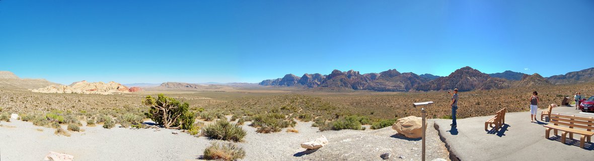 Wide desert landscape showing red rock mountains in the distance, sparse desert vegetation, and a paved visitor overlook with wooden benches and tourists
