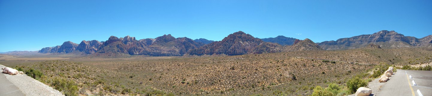 Panoramic view of red sandstone mountains rising above desert scrub vegetation under a clear blue sky, with a paved parking area visible in the foreground