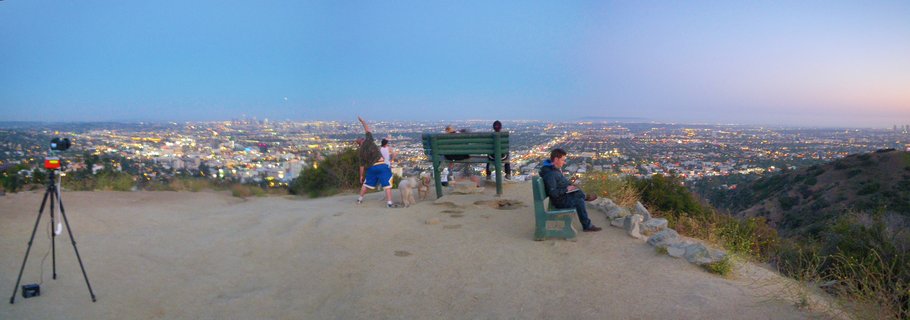 Panoramic twilight view of Los Angeles cityscape from Griffith Observatory viewpoint showing urban sprawl with city lights glowing during blue hour, visitors with dogs and camera tripod on dirt viewing area with green park benches in foreground, deep blue gradient sky above, June 2011