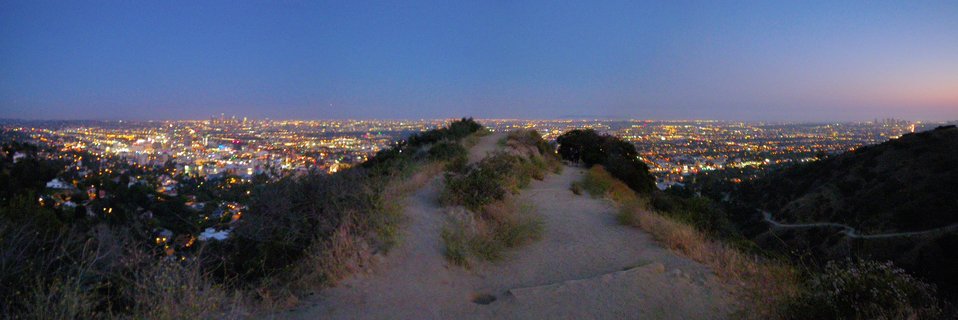 Panoramic view of Los Angeles city lights at dusk from a hilltop dirt trail with twilight sky gradient from blue to pink