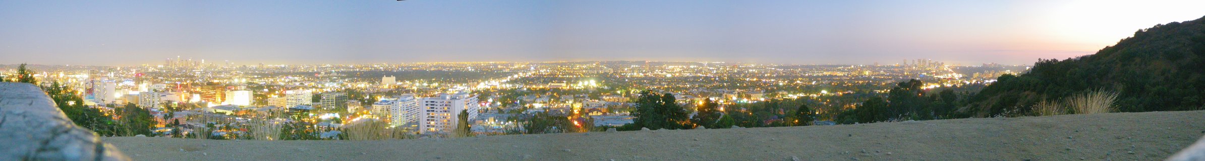 Wide panoramic photograph of Los Angeles city lights at twilight with hills in foreground and urban sprawl extending to horizon under purple-blue evening sky