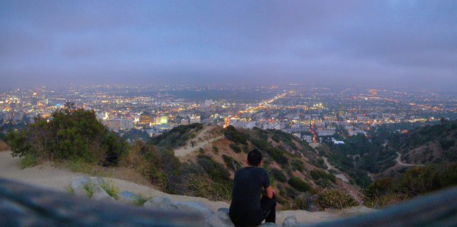 Person sitting on hillside overlooking Los Angeles city lights at dusk with purple-blue twilight sky