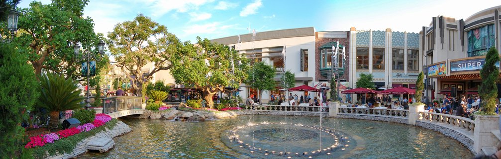 Outdoor entertainment complex with a large circular fountain in the foreground, Art Deco buildings with restaurants, red umbrellas over outdoor dining areas, landscaped gardens with pink flowers and trees, people walking and dining during evening