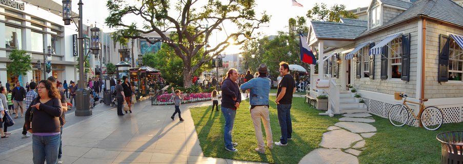 Outdoor shopping center with people walking and standing on grass near a gray shingled cottage with blue striped awnings and a Texas flag, modern retail buildings on the left, large tree with sun filtering through branches