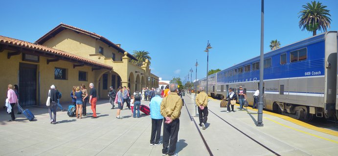 People waiting on a train station platform next to a blue and silver Amtrak Surfliner train at Santa Barbara station with Spanish Colonial architecture and palm trees under clear blue sky