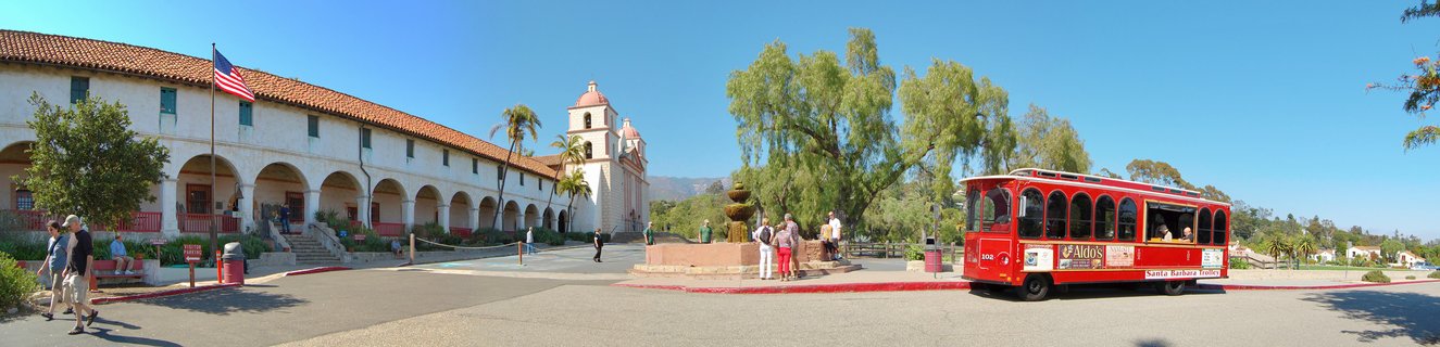 Historic white mission building with red tile roof, twin bell towers, arched colonnade, American flag, fountain, red tourist trolley, and pedestrians under clear blue sky