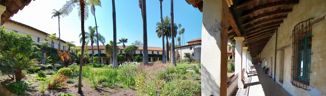 Spanish Colonial courtyard with tall palm trees, flower gardens, and a covered walkway with white columns in Santa Barbara