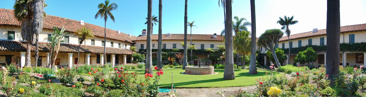 Panoramic view of Santa Barbara Mission courtyard with Spanish Colonial buildings, red tile roofs, tall palm trees, central fountain, green lawn, and rose gardens under blue sky