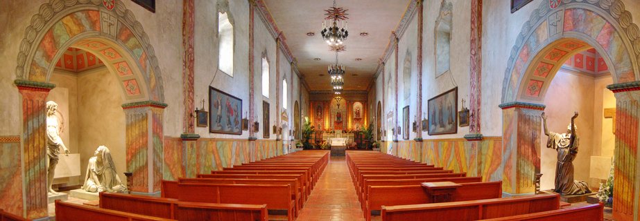 Church interior with colorful painted arches on both sides, rows of wooden pews down the center aisle, chandeliers hanging from ceiling, religious statues in alcoves, and altar visible at the far end