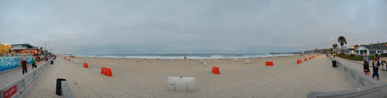 Wide beach scene showing orange barriers on sand with ocean waves, pier in distance, boardwalks with people on both sides, overcast sky