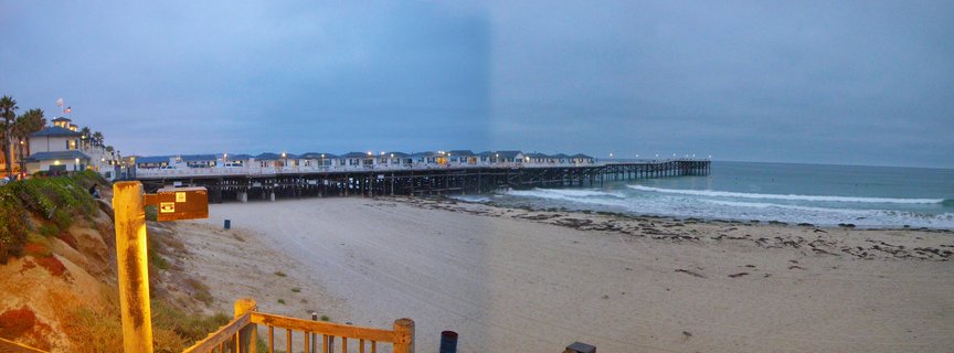 Illuminated wooden pier extending over ocean at dusk with sandy beach, waves, and coastal bluff with palm trees