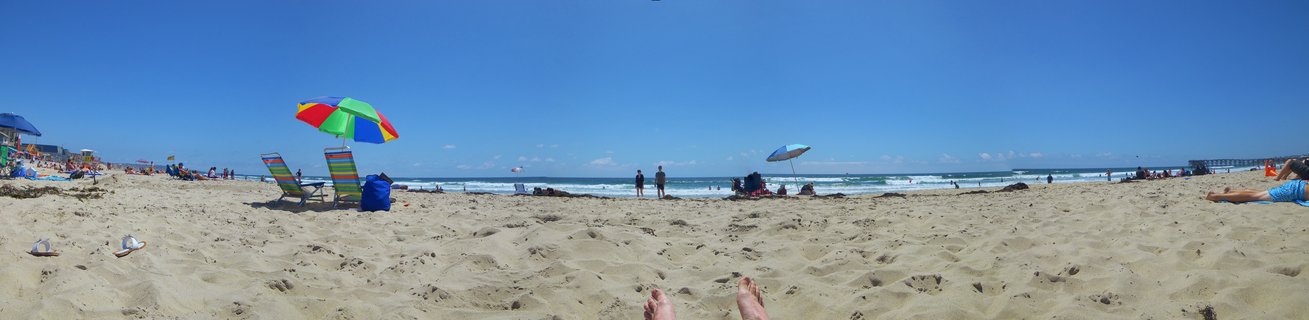 Wide panoramic view of a sandy beach with colorful umbrellas, beach chairs, and people relaxing under a clear blue sky with ocean waves and a pier in the distance