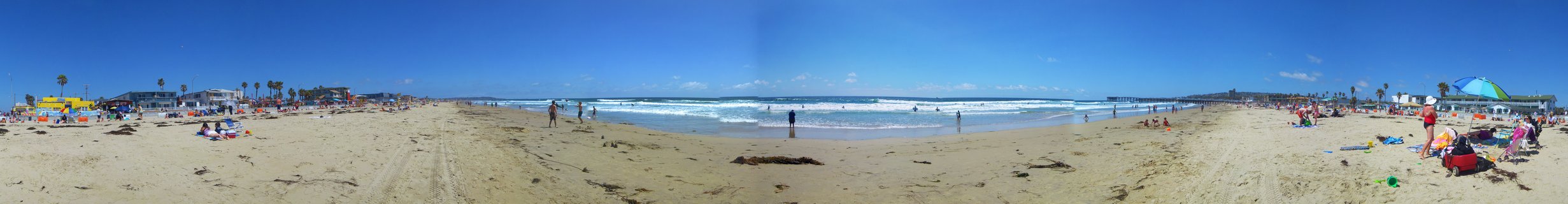 Panoramic photograph of a crowded sandy beach in San Diego with people, beach umbrellas, ocean waves, palm trees, buildings, and a pier under a clear blue sky