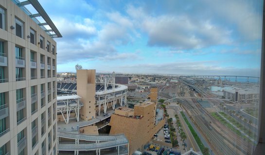 Aerial cityscape view of San Diego showing Petco Park baseball stadium with white steel structure, Coronado Bridge spanning San Diego Bay, urban buildings, train tracks, and infrastructure under blue sky with scattered clouds during late afternoon