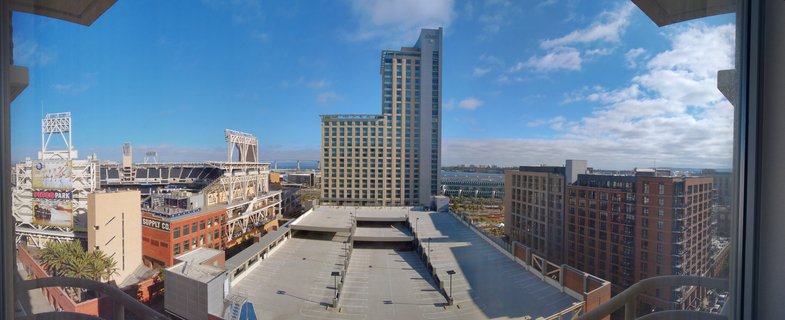 Cityscape photograph of downtown San Diego showing PETCO Park baseball stadium with white steel framework, modern hotel and office buildings, brick warehouse with Supply Co. signage, parking structure, and San Diego Bay with Coronado Bridge visible in the distance under blue sky with clouds