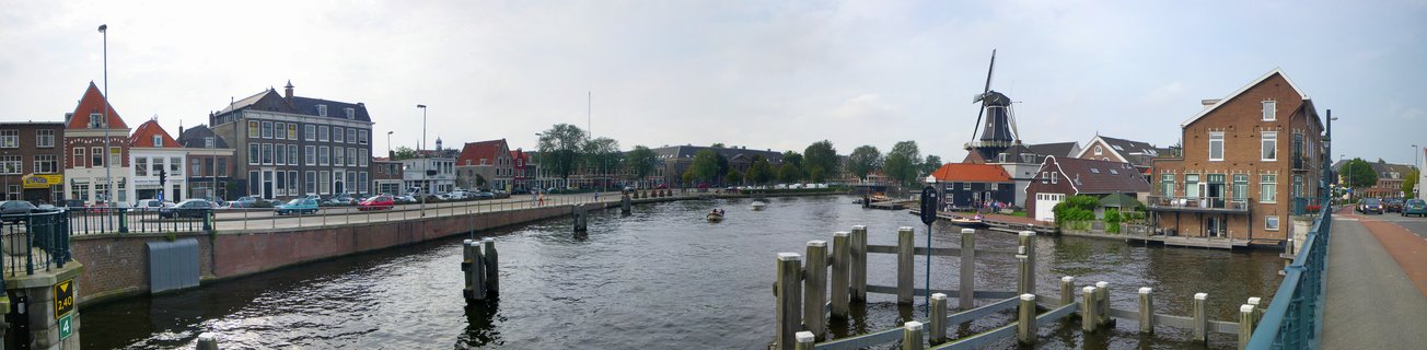 Panoramic view of a canal in Haarlem, Netherlands showing traditional Dutch buildings, a windmill, bridge with wooden posts, and water with reflections under cloudy sky