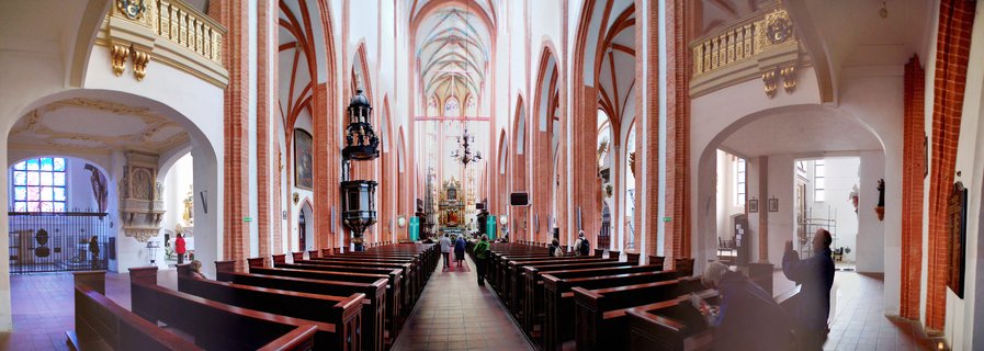 Wide interior view of a Gothic church with red brick pillars, vaulted ceilings, wooden pews, and visitors walking down the center aisle toward an ornate altar