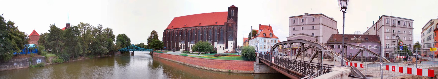 Panoramic view of Wrocław showing a large brick Gothic church with red roof along the Oder River, with metal bridges and historic buildings under an overcast sky